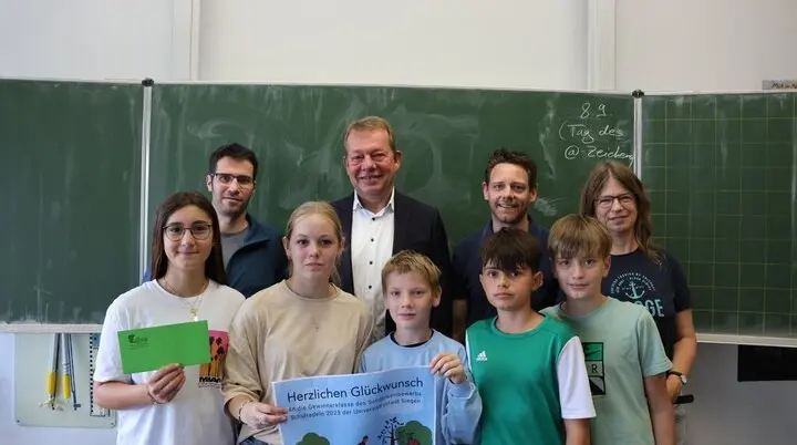Eine Gruppe steht vor einer Tafel in einem Klassenraum, in der hinteren Reihe stehen Erwachsene in der vorderen Reihe 5 Schüler/innen. Die Schüler/innen halten ein Plakat mit der Aufschrift "Herzlichen Glückwunsch" und einen grünen Umschlag in der Hand.  | © Universitätsstadt Siegen