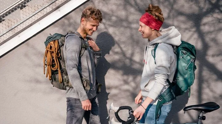 Eine Frau mit Rucksack hält ihr Rad, begegnet dabei einem Mann mit Kletterausrüstung. Er schaut das Rad interessiert an, beide wirken glücklich | © DAV/Marisa Koch