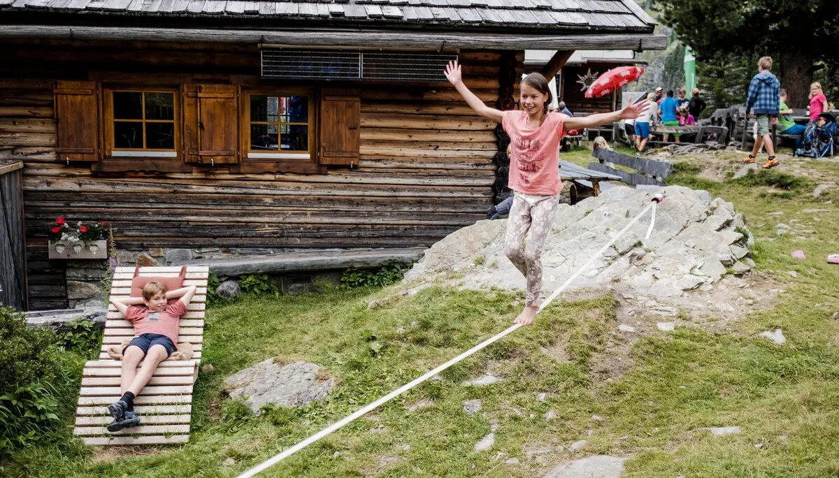 Zwei Kinder balancieren auf einem Seil, das vor einer Berghütte gespannt ist | © DAV/Hans Herbig